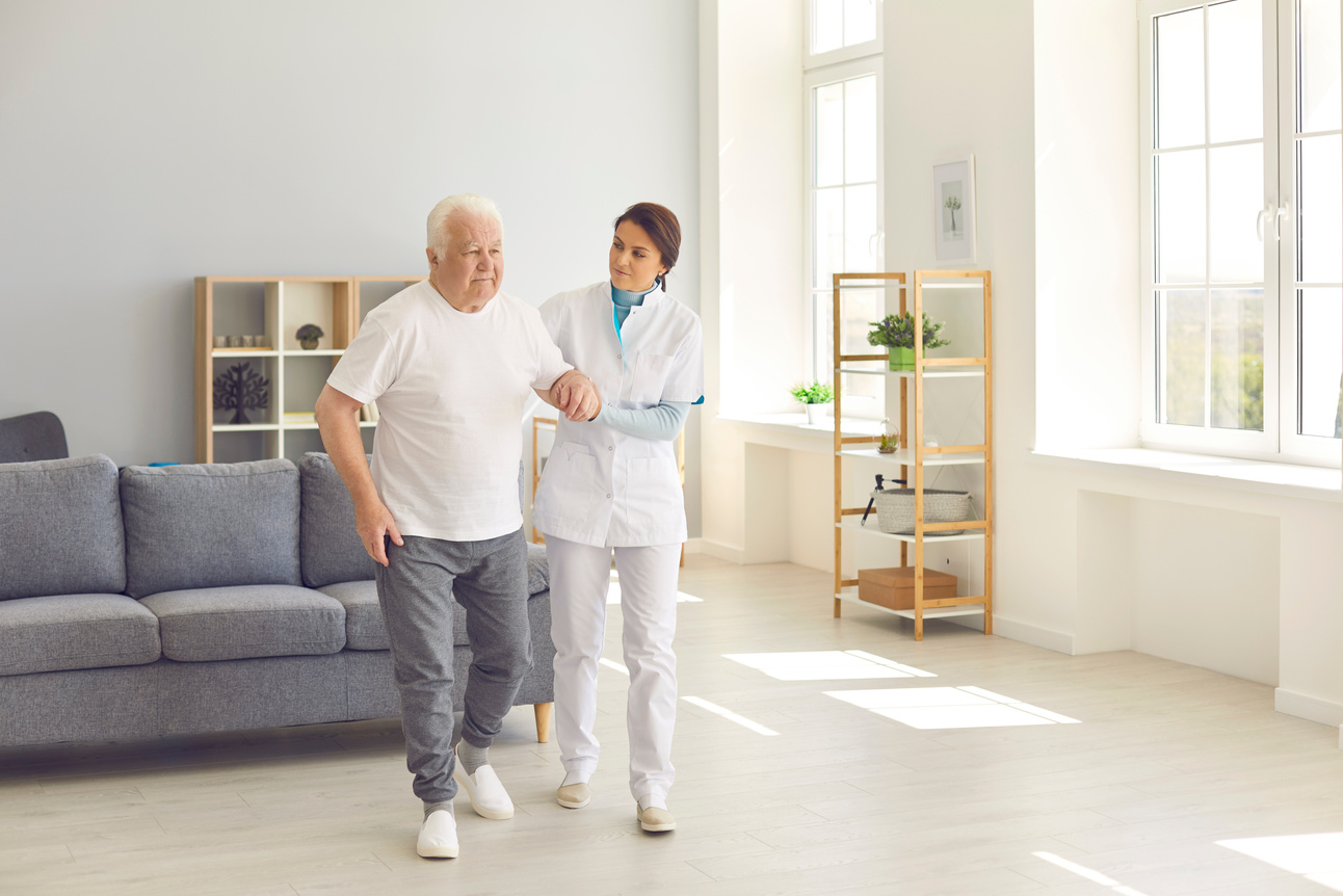 Healthcare Worker Helping Senior Man to Walk in Office of Modern Hospital or Geriatric Rehab Center